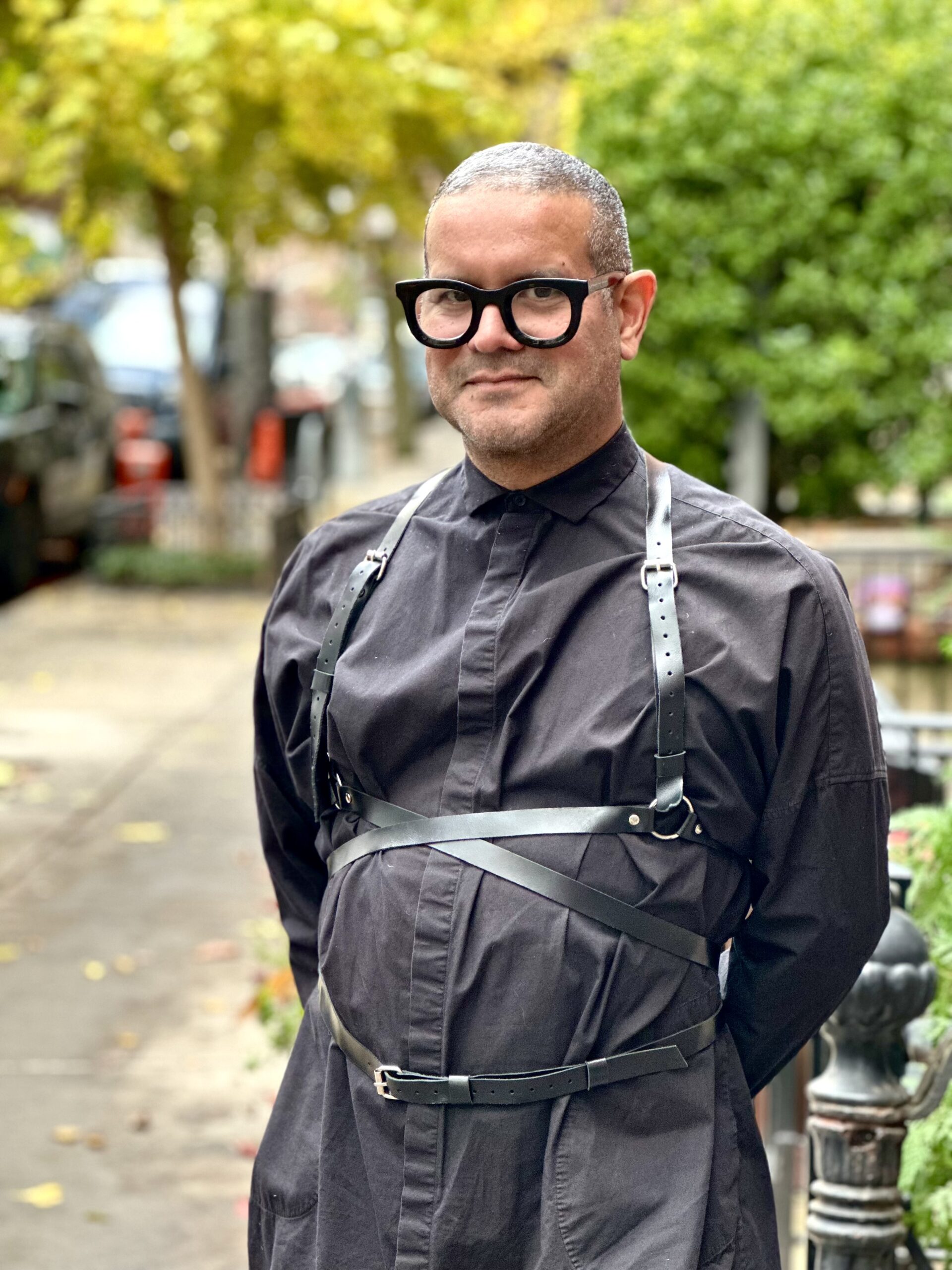 Person in black outfit with leather harness stands on a leafy street.