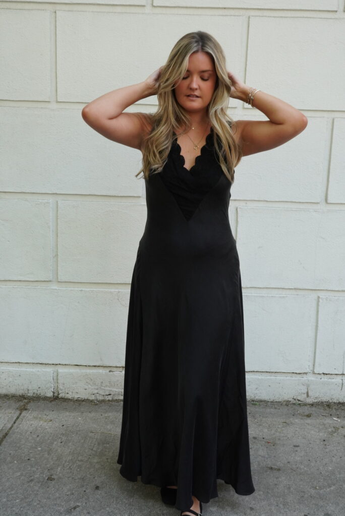 Woman in a black dress stands with hands on head against a white wall.