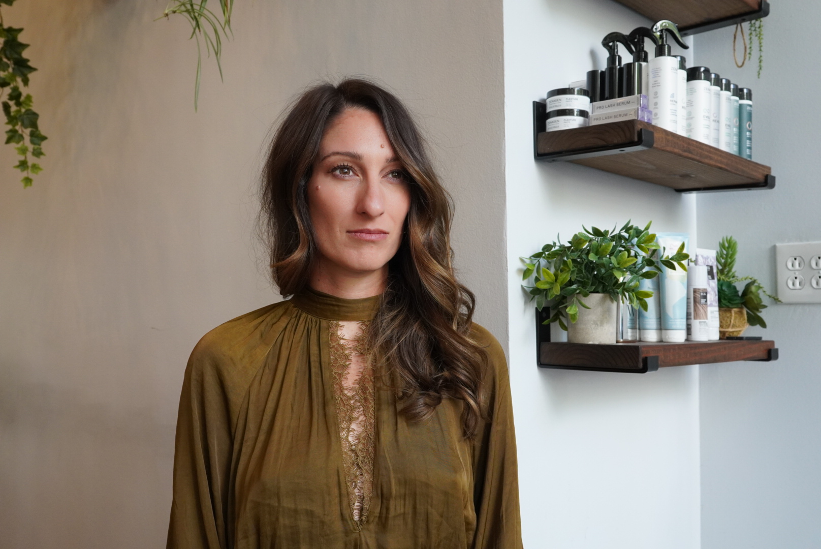 Woman with long hair stands near shelves with beauty products and plants.