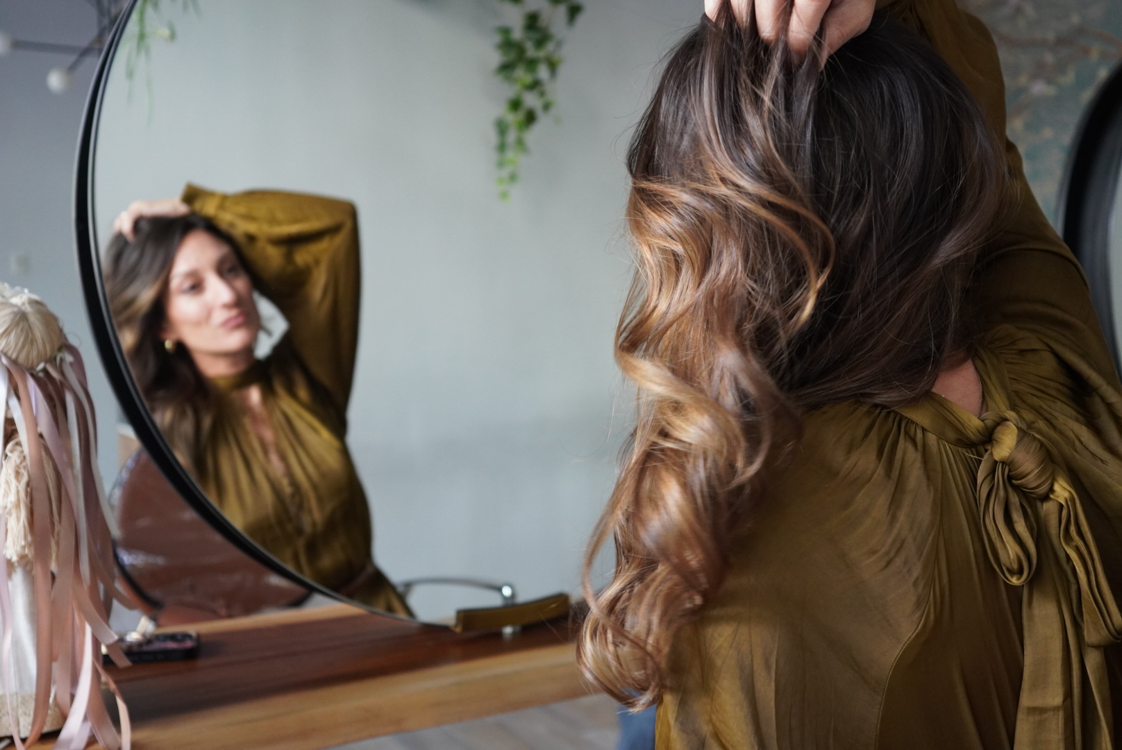 Woman with wavy hair looks in a round mirror while adjusting her hairstyle.