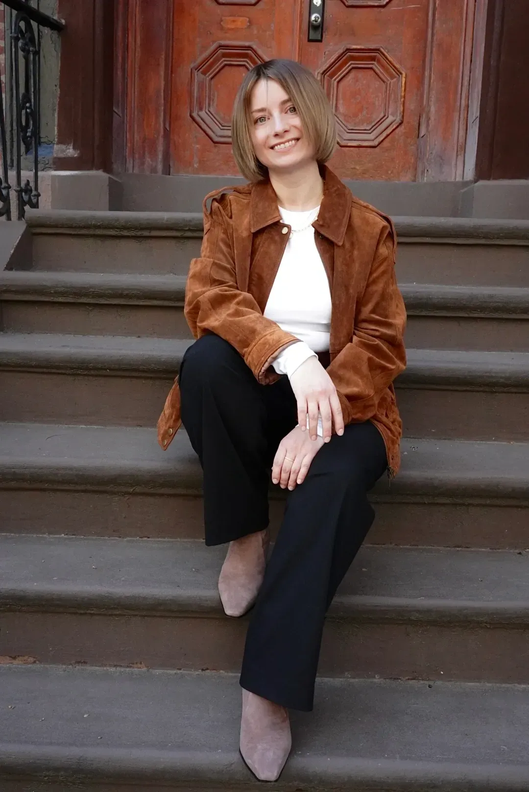 Person sitting on steps wearing a brown jacket and black pants, smiling at the camera.