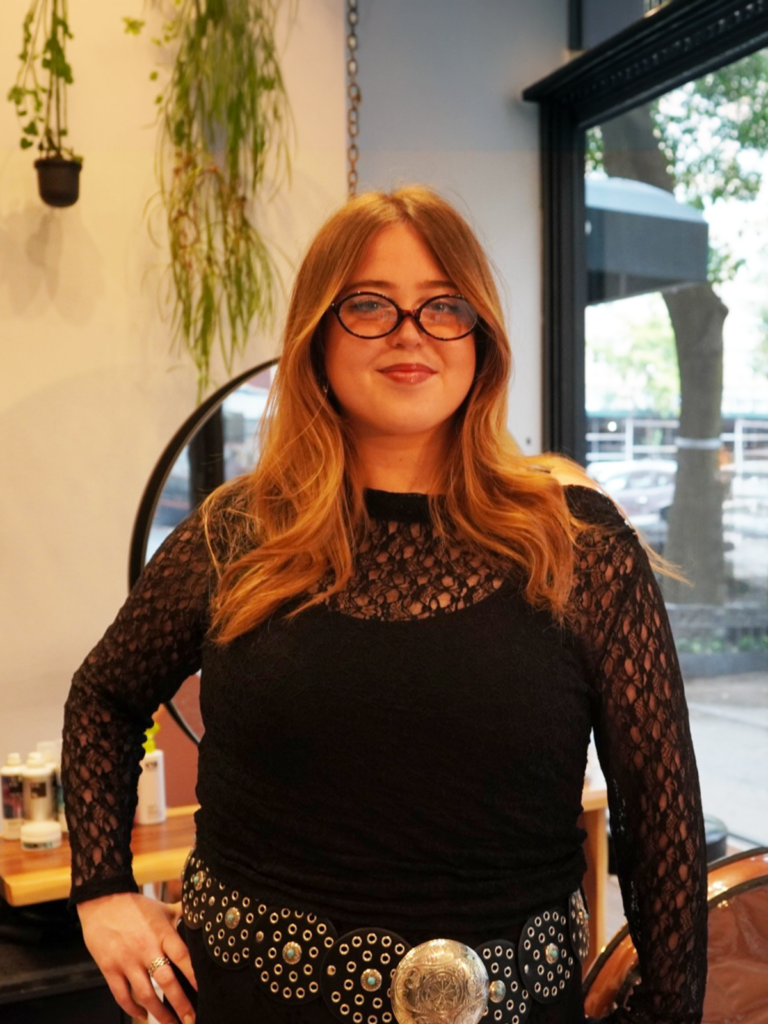 Person with glasses and long hair wearing a black lace outfit, standing indoors.